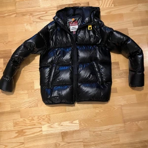 Svart pufferjacka från Parajumpers - Säljer en svart pufferjacka från Parajumpers som aldrig är använd, modell Buzz Puffers. Jackan har glansig finish, huva och dragkedja framtill. Fylld för extra värme och har klassisk pufferlook. Perfekt för kalla dagar och riktigt snygg streetstil.