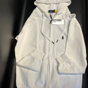 Vit hoodie  - Snygg vit hoodie med dragkedja framtill, huva med snörning och klassisk broderad logga på bröstet. Mjuk bomullskvalitet och ribbade muddar vid ärmslut och nederkant. Perfekt för en clean och stilren look.