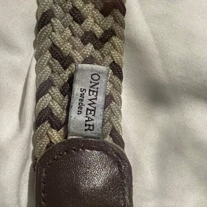 Randigt flätat bälte från ONEWear - Snyggt flätat bälte från ONEWear Sweden med ett randigt mönster i beige och brunt. Bältet har detaljer i brunt skinn och ett silvrigt spänne. Perfekt för att ge din outfit en cool touch. Flätad design gör det flexibelt och bekvämt.