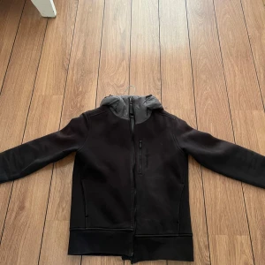 Svart jacka med huva - Snygg svart jacka med grå huva och dragkedja framtill. Den har en stilren design med långa ärmar och en bekväm passform. Säljs pga har blivit för liten, den är i barn storlek m och är lite sol blekt men inget man tänker på