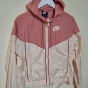 Nike vindjacka rosa och vit M - Nike vindjacka i snygg tvåfärgad design med ljusrosa och vit panel. Jackan har huva, hel dragkedja framtill och elastisk nederkant. Perfekt för dig som gillar sportig stil och vill ha något lätt och bekvämt. Ikonisk Nike-logga på bröstet.