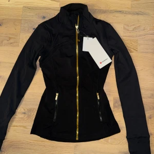 Define Jacket från Lululemon - Snygg svart träningsjacka från Lululemon med guldiga dragkedjor framtill och på fickorna. Jackan har figurnära passform, hög krage och långa ärmar. Perfekt för dig som vill ha en sportig och stilren look. Taggarna sitter kvar.