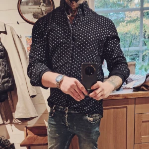 Svart skjorta med vita prickar - Svart premiumskjorta med små vita prickar och klassisk krage. Skjortan har lång ärm och knappar framtill. Passformen är slim och materialet känns som bomull, vilket gör den bekväm och lätt att bära. Perfekt för dig som gillar stilrena detaljer och mönster. Storleken är L men något liten i storleken passar även M. 