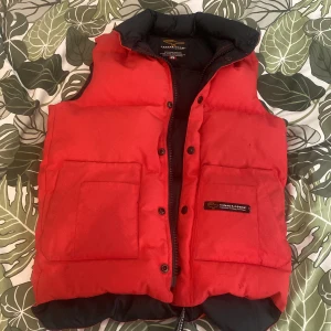Röd dunväst från Canada Goose - Snygg röd dunväst från Canada Goose med svart insida och hög krage. Västen har två stora fickor framtill, knappar och dragkedja. Perfekt för kalla dagar när du vill hålla värmen och ändå se cool ut. Klassisk logga på fickan och quiltad design.