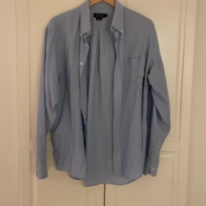 Ljusblå Taylor's skjorta - Snygg ljusblå skjorta från Taylor's Club Collection i storlek L men passar M, Skjortan har klassisk krage, knappar framtill och ett broderat emblem på bröstfickan. 