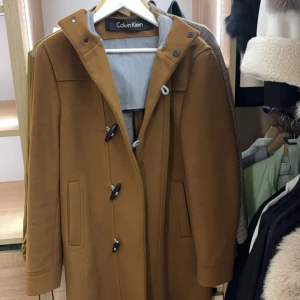  Calvin Klein kappa - Snygg brun duffelcoat från Calvin Klein med huva och klassiska knäppningar framtill. Jackan har raka ärmar, två fickor och är fodrad med grått tyg på insidan. Perfekt för dig som vill ha en stilren och tidlös look. Köpt för 5500kr
