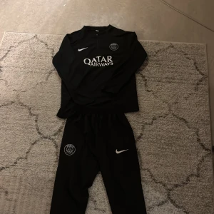 Svart PSG fotbollströja Nike 18 - Svart långärmad fotbollströja från Nike med Paris Saint-Germain-logga på bröstet och Qatar Airways-tryck. Tröjan har en stilren design med vit Nike-swoosh och är gjord i ett lätt, sportigt material som andas. Perfekt för dig som gillar PSG och vill ha en snygg och bekväm tröja.