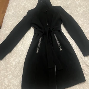 Svart kappa från Vero Moda med bälte - Svart kappa från Vero Moda med hög krage, knytbälte i midjan och två snedställda fickor med dragkedjor. Långärmad och figurnära modell i ullblandning, perfekt för kyliga dagar. Enkel och stilren design som passar till det mesta.