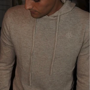 Beige hoodie från Bvaldi med huva - Snygg beige hoodie från Bvaldi med broderad logga på bröstet. Tröjan har huva med snörning och långa ärmar. Mjuk stickad känsla och ribbade muddar vid ärmslut och nederkant. Perfekt för dig som gillar enkel och stilren design.