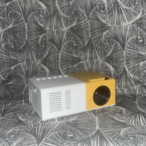 Mini LED Projektor - Kompakt mini LED-projektor i vitt och gult. Utrustad med HDMI, USB och AV-ingångar samt flera kontrollknappar på ovansidan. Perfekt för filmkvällar hemma eller resor. En mindre röd fläck syns på ena sidan, annars i gott skick. Sladden ingår ej.