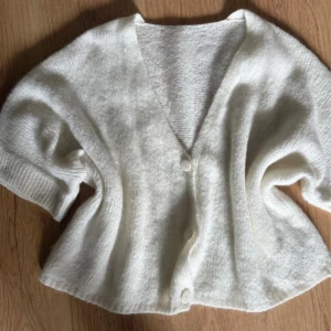 Vit stickad kofta med kort ärm - Mysig vit stickad kofta med korta ärmar och v-ringad hals. Koftan har två knappar framtill och breda ribbade muddar vid ärmslut. Perfekt att slänga över en topp för extra lager och chill vibe.