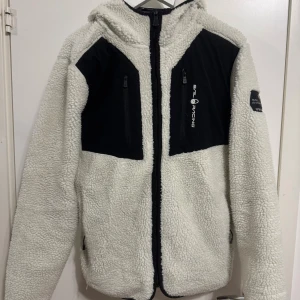 Vit teddyjacka från Sail Racing - Säljer en vit teddyjacka från Sail Racing med svarta detaljer på bröstet och huvan. Jackan har huva, två bröstfickor med dragkedja och broderad logga på ärmen. Materialet är mjuk fleece och jackan har hel dragkedja framtill. St M passar till S också  använd ca 3 gånger är som ny ! Nypris är 2999kr mitt pris 700kr