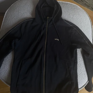 Svart hoodie från BOSS med dragkedja - Svart hoodie från BOSS med guldfärgad broderad logga på bröstet. Tröjan har hel dragkedja, huva med snörning och diskreta kontrastsömmar längs axlarna. Perfekt för dig som gillar stilren streetwear med premiumkänsla.