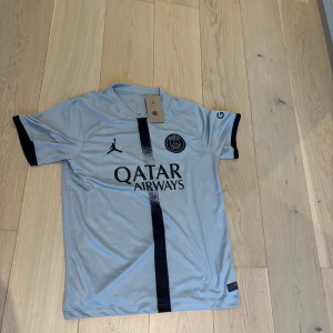 PSG fotbollströja Jordan x Qatar - Snygg ljusblå PSG fotbollströja med svart vertikal rand och svarta detaljer på ärmsluten. Tröjan har korta ärmar, Paris Saint-Germain-logga på bröstet och Jordan-logga. Tillverkad i lätt och ventilerande polyester, perfekt för match eller träning.