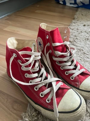Röda Converse Chuck Taylor High - Säljer ett par klassiska röda Converse Chuck Taylor All Star high tops med vit gummisula och vita snören. Skorna har rund tå, textilmaterial och den ikoniska loggan på sidan. Perfekt för dig som gillar retrostil och vill sticka ut med färg.