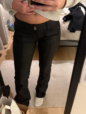 Svarta skinnbyxor med bootcut - Säljer ett par svarta skinnbyxor med bootcut och låg midja. Byxorna har flera fickor framtill med lock och knappdetaljer, samt klassisk dragkedja och knappstängning. Materialet är mjukt och har en lätt stretch för skön passform.