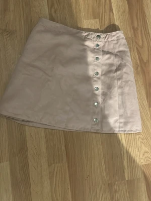 Beige kortkjol med knappar - Säljer en beige kortkjol med snygga silverfärgade knappar framtill. Kjolen har en lätt A-linjeform och är gjord i ett mjukt material som känns skönt mot huden. Perfekt att matcha med en stickad tröja eller t-shirt för en chill look.