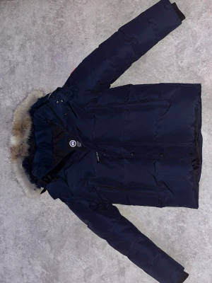 Mörkblå Canada Goose dunjacka med päls - Säljer en mörkblå dunjacka från Canada Goose med äkta päls på huvan och klassisk logga på ärmen. Jackan har dragkedja och knappar framtill, flera fickor och en varm, quiltad insida. Perfekt för kalla vinterdagar och riktigt snygg stil. USAs storlek så den motsvarar S EU. Kvitto finns