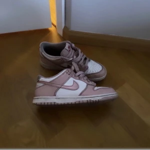 Nike Dunk Low i rosa och vitt - Säljer ett par Nike Dunk Low sneakers i en snygg kombination av ljusrosa och vitt. Skorna har klassisk snörning, rund tå och platt sula. Perfekt för dig som gillar streetwear och vill ha en stilren sneaker med låg profil.