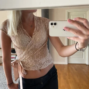 Jättesnygg festtopp! - Säljer en superfin beige omlottopp i spets med korta ärmar och v-ringning. Toppen knyts i sidan med ett sidenband som ger en snygg rosettdetalj. Perfekt för dig som gillar trendig stil och vill ha något extra till outfiten. Storlek S!💗