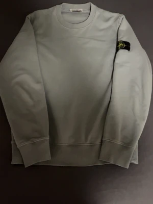 Ljusblå sweatshirt från Stone Island - Snygg grå sweatshirt från Stone Island med klassisk rund hals och ribbade muddar. Tröjan har den ikoniska Stone Island-loggan på vänster ärm och är tillverkad i mjuk bomull. Perfekt för dig som gillar stilrena och enkla plagg med streetkänsla.