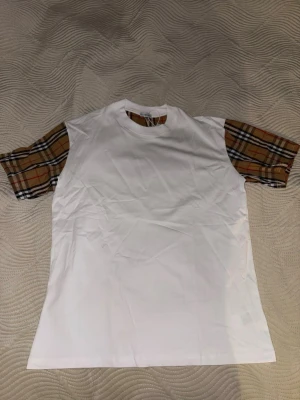 Vit Burberry t-shirt med rutiga ärmar - Säljer en clean vit t-shirt från Burberry med klassiska rutiga ärmar i beige, svart och rött. T-shirten har rund hals och är tillverkad i mjuk bomull. Perfekt för dig som vill ha en stilren look med en touch av lyxig streetstyle.