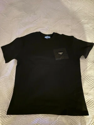 Svart t-shirt från Prada med ficka - Svart t-shirt från Prada med klassisk passform och korta ärmar. På bröstet finns en snygg ficka i nylon med Pradas ikoniska metallogo. Perfekt för dig som gillar stilrena och exklusiva plagg.