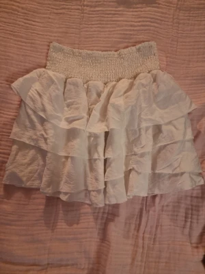 Vit volangkjol med resårmidja - Supersöt vit kortkjol med flera lager volanger och bred smockresår i midjan. Kjolen har en luftig och flowy känsla, perfekt för varma dagar. Materialet känns lätt och skönt mot huden.