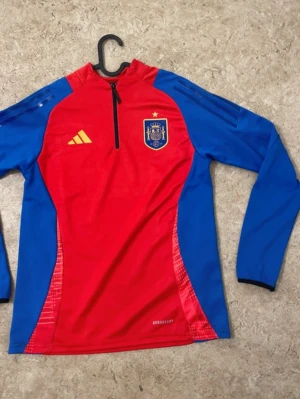 Ingen sko på bilden - Bilden visar ingen sko, utan en långärmad träningströja från Adidas i rött och blått med Spaniens landslagsmärke. Ingen skorelaterad information kan ges.