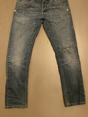 Blå jeans från Dondup med slitningar - Snygga blå jeans från Dondup med slitningar och lite trasig på ena benet. Klassisk femficksmodell med raka ben och sömmar på bakfickorna. Storlek 31