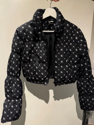 Svart croppad pufferjacka  - Säljer en svart croppad pufferjacka med vit logomönster över hela jackan. Jackan har quiltad design, dragkedja framtill och höga krage. Perfekt för dig som vill ha en trendig och varm look i vinter.