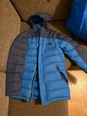 Blå dunjacka från The North Face - Säljer en blå och mörkgrå dunjacka från The North Face med huva och dragkedja. Jackan har två sidofickor med dragkedja och är quiltad för extra värme. Perfekt för kalla dagar och har en snygg, sportig look. Obs dragkedjan behövs fixas hos skräddaren