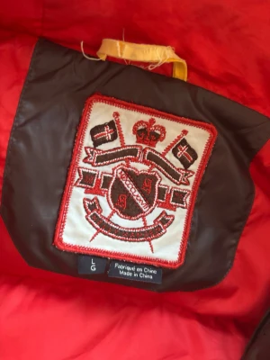 Brun pufferjacka från Helly Hansen - Säljer en brun pufferjacka från Helly Hansen med röd insida och röda detaljer. Jackan har två stora fickor med knappar, hög krage och synligt HH-logga på bröstet. Materialet är glansigt och känns riktigt robust, perfekt för kalla dagar.