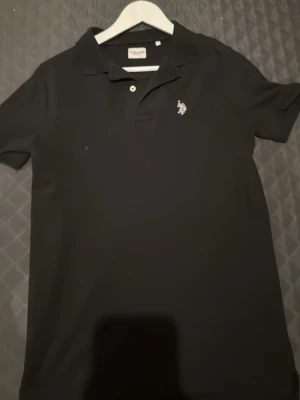 Svart pikétröja från U.S. Polo Assn. - Säljer en klassisk svart pikétröja från U.S. Polo Assn. med korta ärmar och krage. Tröjan har två knappar framtill och en liten broderad logga på bröstet. Perfekt för en clean och sportig look.