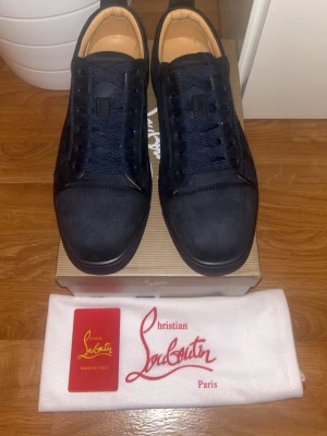 Svarta sneakers från Christian Louboutin - Snygga svarta sneakers från Christian Louboutin med klassisk röd sula och rund tå. Skorna har snörning och är tillverkade i mocka med detaljer i skinn. Perfekt för dig som gillar exklusiva märken och vill sticka ut med en lyxig touch.