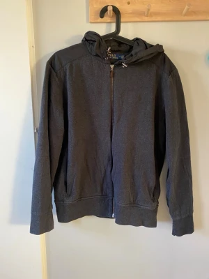  Polo Ralph Lauren cardigan - Grå cardigan från Polo Ralph Lauren med huva och snörning. Jackan har dragkedja framtill och på fickorna, samt detaljer i syntetmaterial på axlar och huva. Stilren design med diskret logga på bröstet. Perfekt för dig som gillar sportig och clean look. Skicket är väldigt bra, utan defekter. Kontakta mig om frågor! 😊