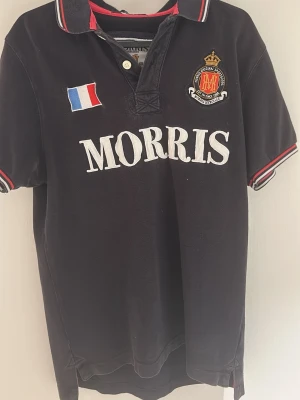 Svart piké från Morris med broderi - En sällsynt pikétröja från Morris med broderat franskt flaggmärke och emblem på bröstet. Stora vita bokstäver framtill, röd och vit rand på krage och ärmslut. Klassisk pikékrage med knappslå, kortärmad och tillverkad i bomull. Den är i toppskick då den knappt är använd. Säljer pga den är lite liten på mig. 