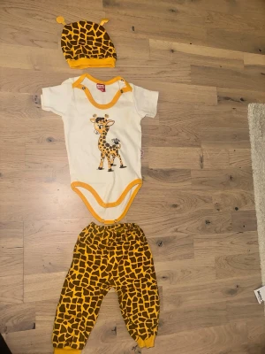 Gul och brun giraffmönstrad babyväst Giraff Set - Body, Byxor och Hatt - Säljer en söt babyväst med giraffmönster i gul och brun från Çikibom. Västen har rund hals, tryckknappar vid axlarna och är gjord i mjuk bomull. Perfekt för dig som gillar djurprint och vill ha något unikt till din lilla.Sött set med en body, byxor och en hatt. Bodyn är vit med en giraff på framsidan och gula detaljer. Byxorna och hatten har ett giraffmönster i gult och brunt.