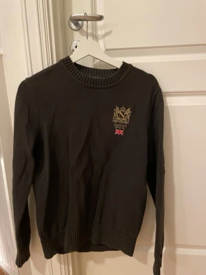 Mörkgrön stickad tröja från Morris - Snygg mörkgrön stickad tröja från Morris med broderad logga på bröstet. Rund hals och ribbade muddar vid ärmslut och nederkant. Perfekt för dig som behöver en varm tröja till vintern.