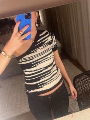 Svartvit oneshoulder topp  - Säljer en cool stickad topp från Lindex i storlek S. Den är svart och vit med ett grafiskt randigt mönster och har oneshoulder-design med kort ärm på ena sidan. Materialet är mjukt stickat och toppen har ribbad kant nedtill för extra passform.