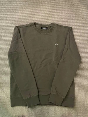 Olivgrön sweatshirt från J.Lindeberg - Snygg olivgrön sweatshirt från J.Lindeberg i storlek M. Tröjan har rund hals, ribbade muddar och ett litet vitt JL-logo tryck på bröstet. Tillverkad i mjuk bomull som är skön att ha på sig. Perfekt för chill dagar och enkel att matcha med jeans eller joggers.