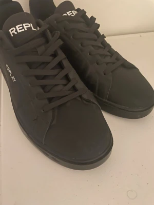 Svarta sneakers från Replay  - Säljer ett par svarta sneakers från Replay med stilren design och vit logga på plösen och sidan. Skorna har rund tå, platt sula och snörning. Materialet är syntet och de passar perfekt till en modern streetstil.