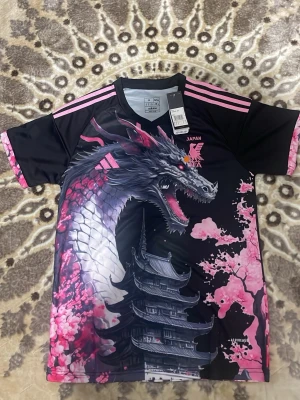 Adidas Japan tröja med drakmotiv M - Säljer en Adidas Japan tröja i slim fit med ett fett drakmotiv, körsbärsblommor och pagoda. Svart bas med rosa detaljer och mönster, samt rosa Adidas-logga och JFA-emblem. Materialet är polyester och tröjan har Aeroready-teknologi för extra komfort.