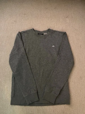 Grå sweatshirt från J.Lindeberg - Säljer en stilren grå sweatshirt från J.Lindeberg i storlek M. Tröjan har rund hals, lång ärm och ett litet vitt logotryck på bröstet. Tillverkad i mjuk bomull som är skön mot huden. Perfekt för dig som gillar enkel och clean design.