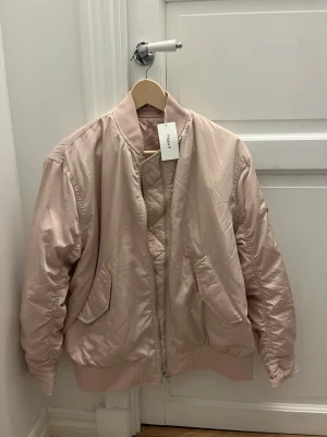 Blushrosa bomberjacka från Filippa K - Snygg puderrosa bomberjacka från Filippa K med klassisk ribbad krage och muddar. Jackan har två fickor framtill och stängs med dragkedja. Helt ny! 