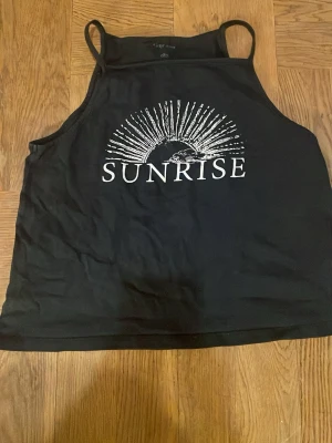 Svart linnetopp med SUNRISE från Ellos - Cool svart linnetopp från Ellos med tryck i vitt som föreställer en soluppgång och texten SUNRISE på bröstet. Toppen har smala axelband och är ärmlös, perfekt för varma dagar. Materialet är mjuk bomull som känns skönt mot huden. Det finns två stycken, när du köper får du båda två. Väl använd. Säljer för att dem blivit för liten.