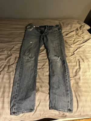 Levi's 501 ljusblå jeans med slitningar - Säljer ett par klassiska Levi's 501 jeans i ljusblå tvätt med snygga slitningar och hål på knäna. Jeansen har rak passform, fem fickor och den ikoniska läderpatchen bak i midjan. W29 L30