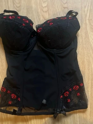 Korsett topp med röda broderade rosor - Säljer en svart korsett topp med vadderade kupor och justerbara axelband. Dekorerad med broderade röda rosor och spetsdetalier. Stängs med hyskor i ryggen. Jag är 75b och kuporna är för stora för mej. Perfekt till att skapa en snygg siluett💕