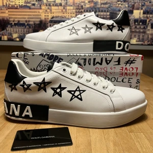 Dolce & Gabbana vita sneakers med stjärnor - Säljer ett par vita sneakers från Dolce & Gabbana med svart text och stjärnmönster på sidorna. Skorna har rund tå, platt sula och svart hälparti. Materialet är skinn och de har snörning framtill. Boxen har coolt grafiskt tryck och autenticitetskort medföljer.