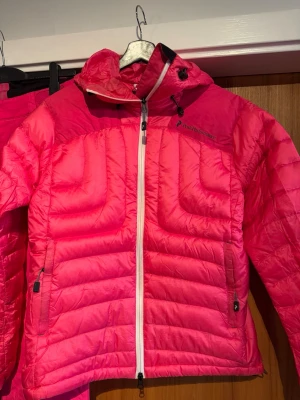 Peak performance Rosa Dun pufferjacka  L - Säljer en riktigt snygg rosa Dun pufferjacka från Peak Performance i storlek L. Jackan har huva, vita dragkedjor och två sidofickor med dragkedja. Perfekt för kalla dagar och ger en sportig vibe. Materialet är lätt och quiltat för extra värme.